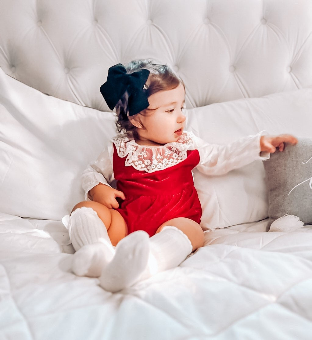 baby wearing the Baby Girl Vintage Lace Red Velvet - White Sleeves Romper. with a dark green bow in hair and white knee high socks.