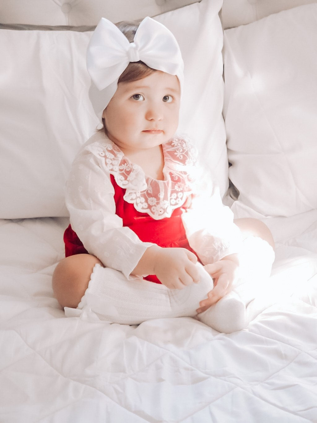 child wearing the Baby Girl Vintage Lace Red Velvet - White Sleeves Romper. Also has white boy headband on head, and white knee high socks.