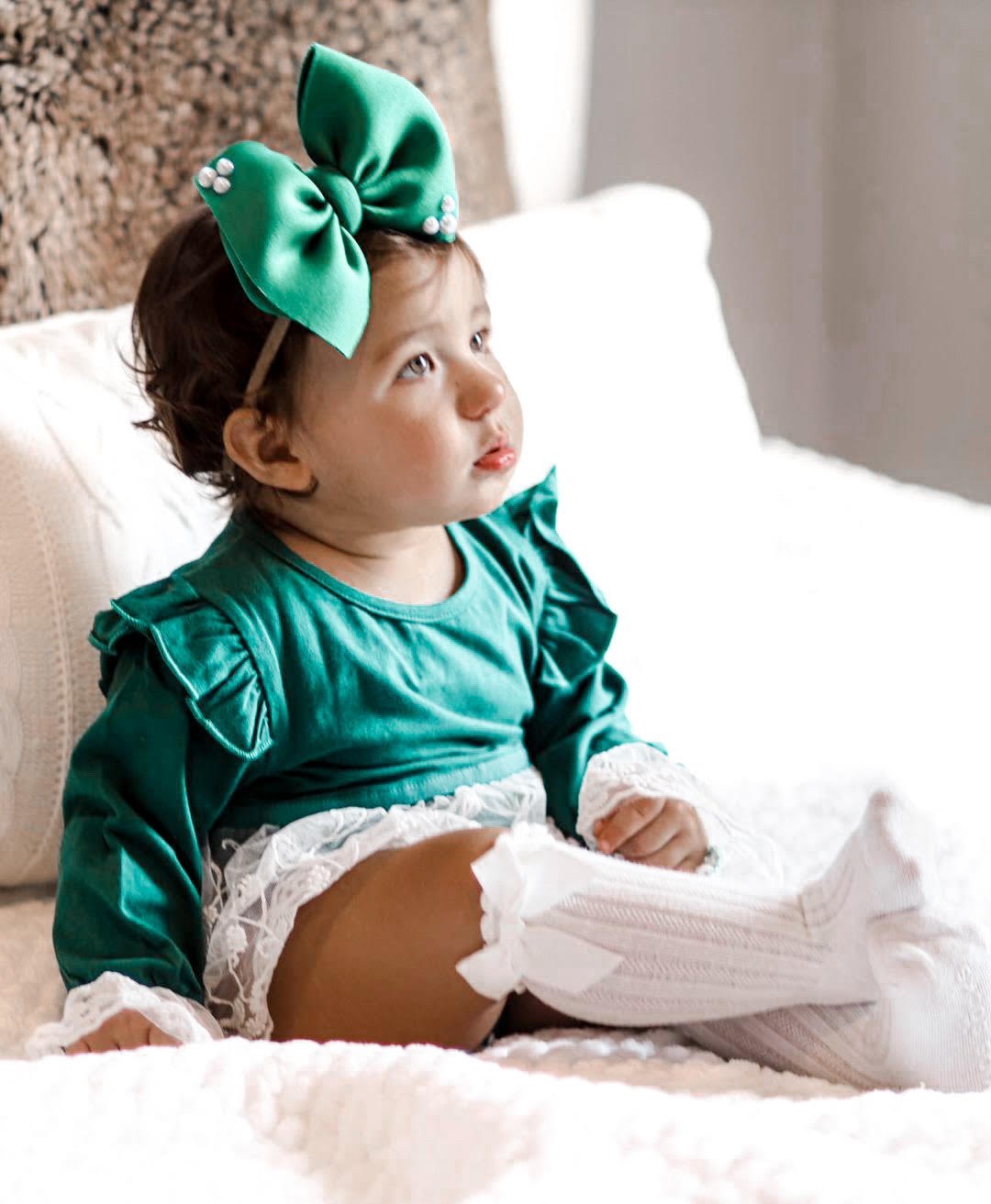 child wearing the Baby Girl Vintage Lace Green Lace Skirted Romper. Also wearing a green bow & button headband, as well as white knee high socks.