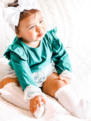child wearing the Baby Girl Vintage Lace Green Lace Skirted Romper. Also wearing a white bow headband & white knee high socks.