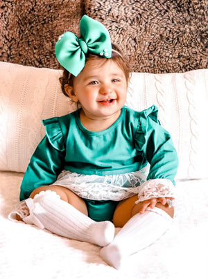 child wearing the Baby Girl Vintage Lace Green Lace Skirted Romper. Also wearing a green bow & button headband, as well as white knee high socks.