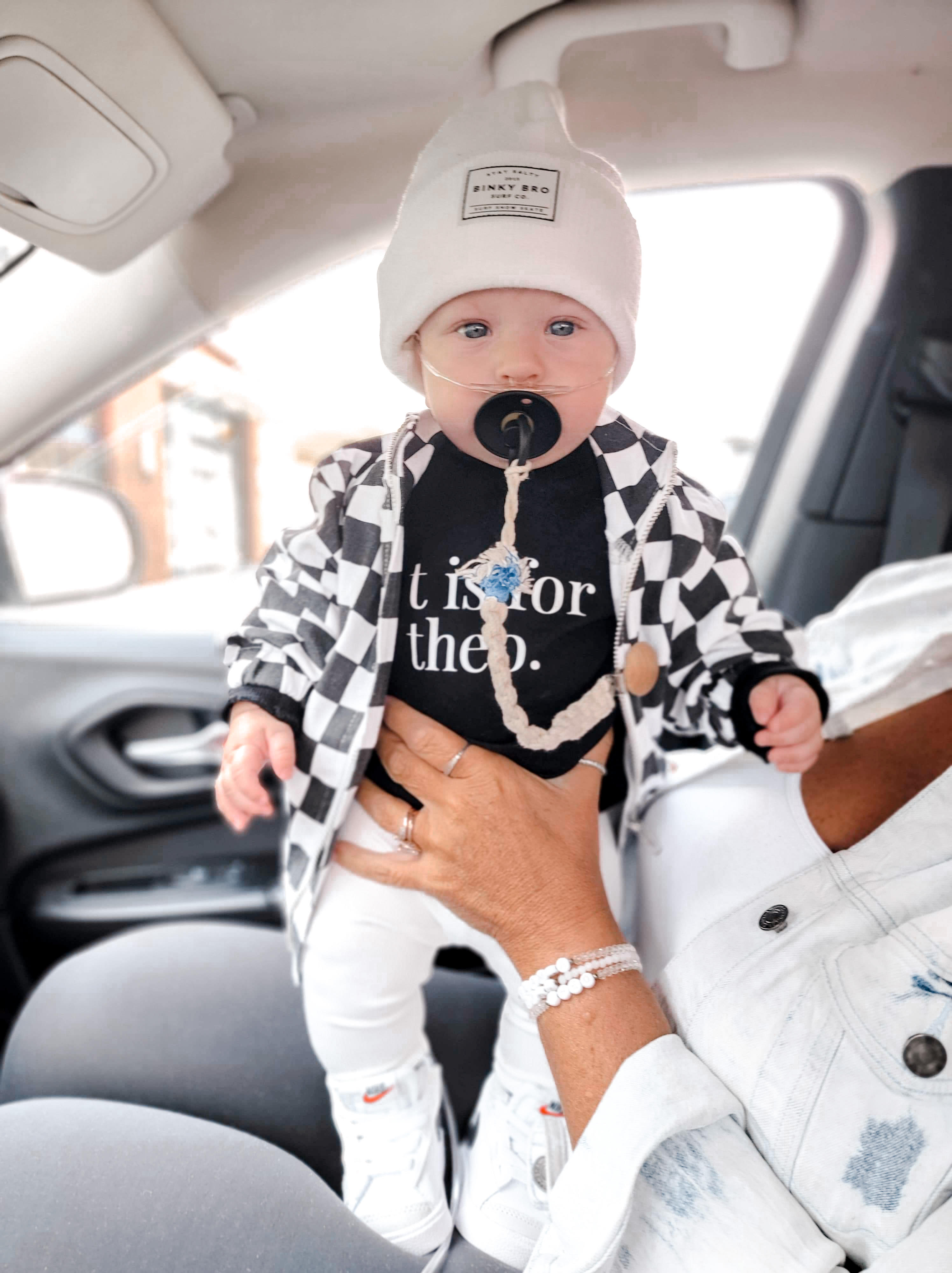 child in black and white check/block jacket, white winter hat, and black soother