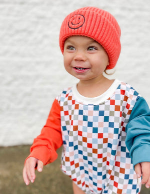 Smiling boy in Salmon color smiley hat and Theo & Me Multi Color Checkers Bubble Romper. 1 Reddish Sleeve, 1 blueish sleeve.