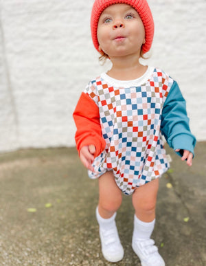 Curious boy in Salmon color smiley hat and Theo & Me Multi Color Checkers Bubble Romper. 1 Reddish Sleeve, 1 blueish sleeve.