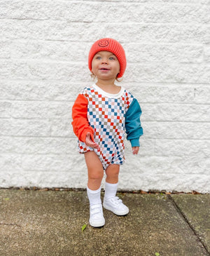 Smiling boy against a white brick wall in Salmon color smiley hat and Theo & Me Multi Color Checkers Bubble Romper. 1 Reddish Sleeve, 1 blueish sleeve.