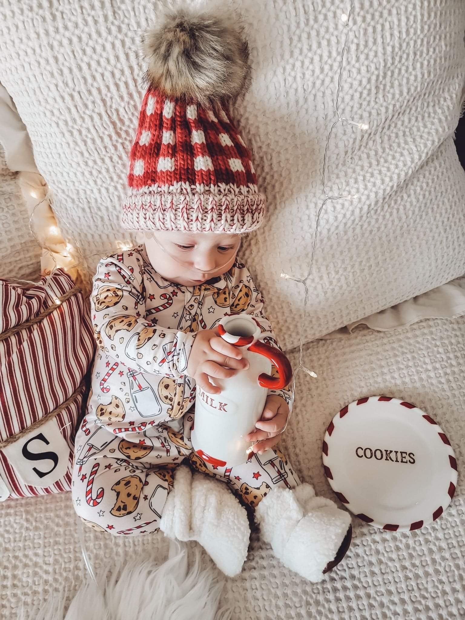 kid wearing the Holiday Zippies Sleepers - Milk & Cookies with red check knit hat & white soft shoes
